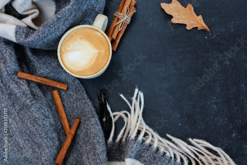 Cup of cappuccino, with cinnamon, warm gray scarf and dry fall leaf on black background. Autumn hot drink, cozy coffee shop mood concept