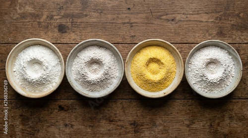 Top-down photograph of four neat mounds of flour in shallow ceramic bowls arranged in a row on a rustic wooden surface