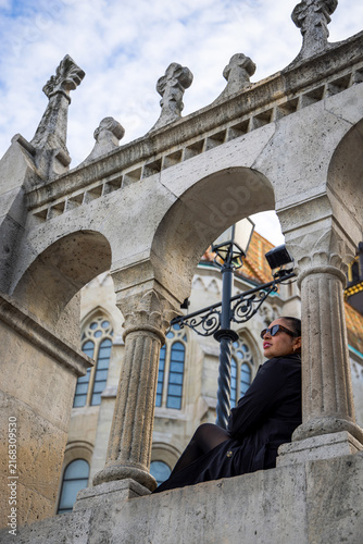 Принти на полотні An elegant woman in a black outfit relaxing outdoors, seated against a historic stone architectural structure with ornate arches