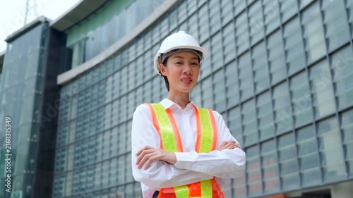 Smiling Asian woman civil engineer wearing hard hat standing with arms crossed in front of glass office building. Professional female engineering career concept.