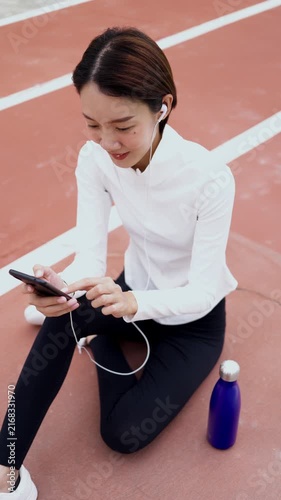 Young Asian woman resting on sports running track with water bottle, using mobile phone with earphones. Active healthy lifestyle, fitness app, hydration, and wellness concept. Vertical Shot