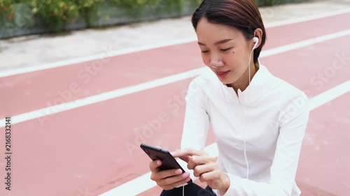 Young Asian woman resting on sports running track with water bottle, using mobile phone with earphones. Active healthy lifestyle, fitness app, hydration, and wellness concept.