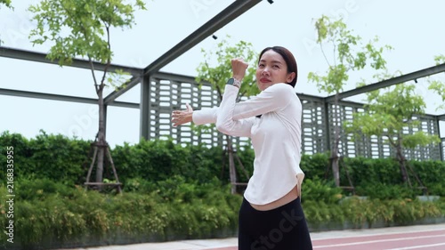 Young Asian sportswoman doing arm stretching exercise on red running track in outdoor park. Workout preparation, cool down stretch, fitness training, and wellness concept.