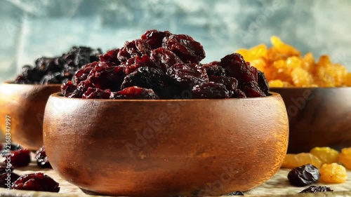 Low angle rotating display of three raisin varieties in wooden bowls with scattered dried fruit on a rustic surface
