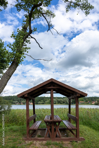 Empty wooden picnic shelter with benches and a table beside a reed-lined lake under a bright blue sky with large summer clouds.