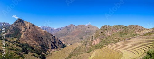 View of the ancient Inca agricultural terraces at the Pisac archaeological site, set against rugged Andean mountains under a clear blue sky in Pisac, Cusco, Peru.