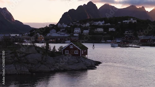 Traditional wooden rorbu cabin stands on rocky shore overlooking coastal bay and mountainous peaks in Reine, Lofoten, Norway at dawn.
