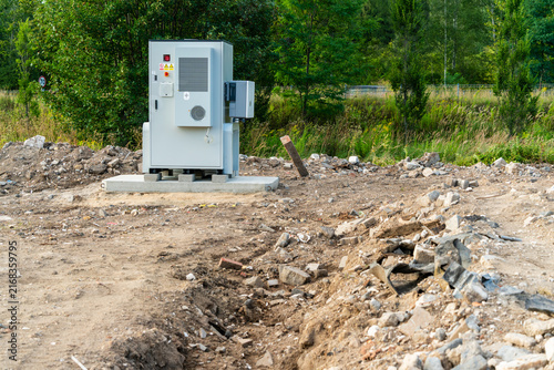 View of a modern electrical control cabinet on a concrete base in an undeveloped area with scattered debris and trees in the background in Dabrowa Gornicza, Silesian Voivodeship, Poland.
