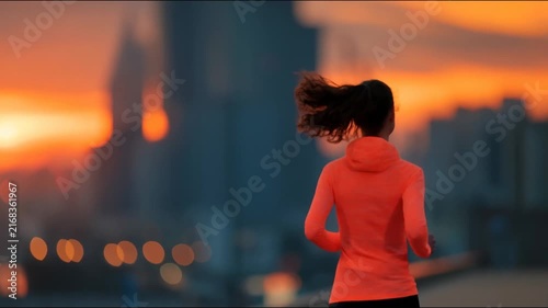 A woman in an orange hoodie runs away from the camera against a blurred city sunset with bokeh lights.