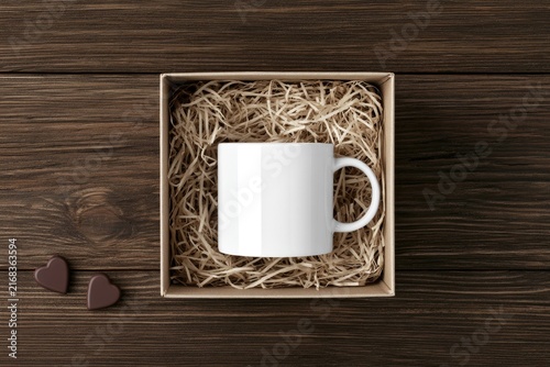 White mug nestled in straw-filled box with heart candies on wood