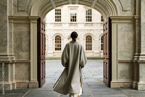 Elegant woman in long grey coat walking through marble archway toward neoclassical courtyard building.