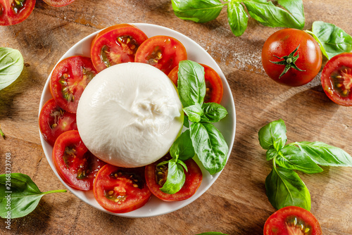 Flat lay of burrata cheese surrounded by cherry tomatoes and basil on wooden table.