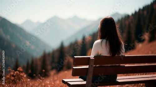 Young woman with long hair sitting on a wooden bench admiring majestic mountain peaks during autumn season.