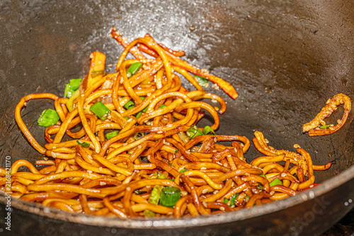 Action shot of a culinary chef cooking savory Singapore noodles with vegetables and protein in a commercial kitchen.