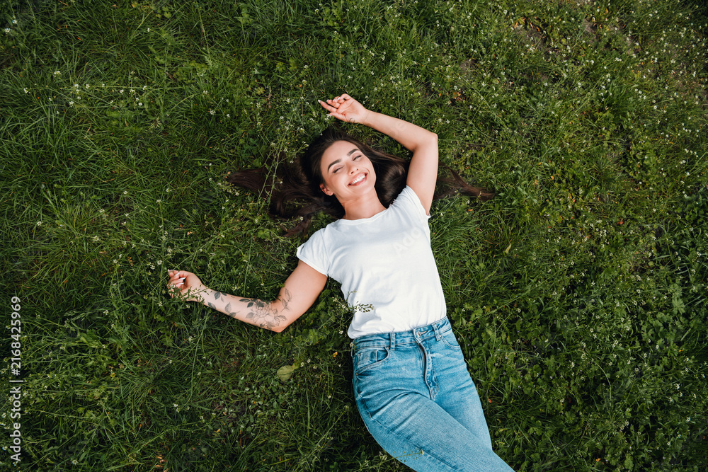 Fototapeta premium Young woman relaxing on green grass, smiling and enjoying a sunny outdoor moment with casual jeans and white t-shirt, showing a floral arm tattoo and carefree summer vibe