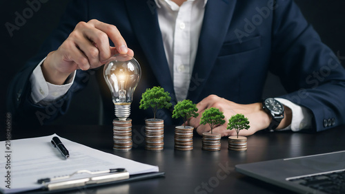 Business professional lighting a bulb surrounded by growing money trees and coins on a dark table