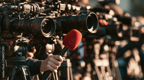 A hand holding a red microphone in front of a row of professional video cameras at a press conference.