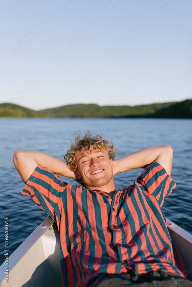 Fototapeta premium Smiling young man reclining on a boat bow with arms behind head at a lake in striped shirt for summer portrait background and travel poster