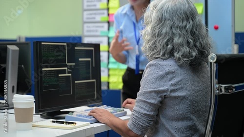 Senior Asian female software engineer coding on a desktop computer while a team member manages project tasks on an agile scrum board