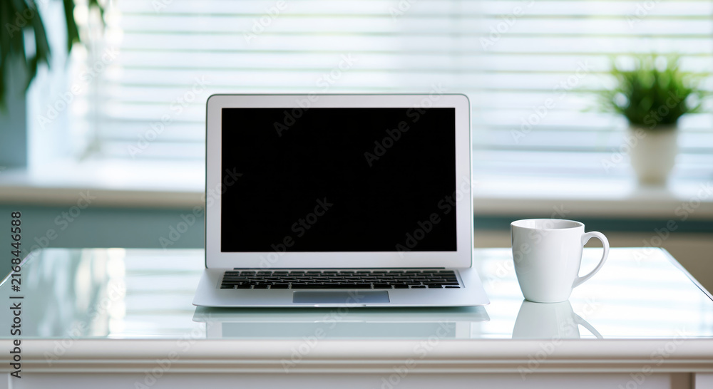 Fototapeta premium Modern silver laptop and white coffee cup on glass desk in bright home office with window blinds and plant, minimalist workspace with calm atmosphere