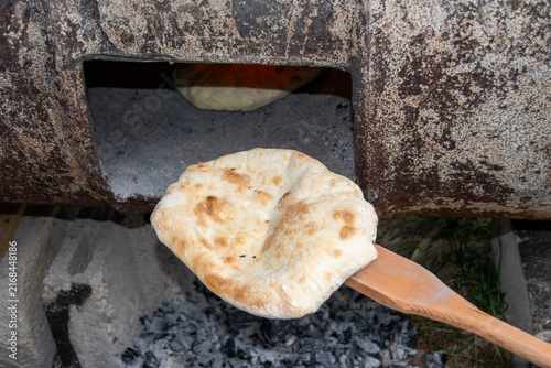 Traditional bread baking inside a rustic wood-fired barrel oven next to burning logs and hot embers. Authentic outdoor baking process in a rural village setting.