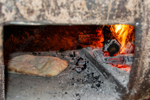 Traditional bread baking inside a rustic wood-fired barrel oven next to burning logs and hot embers. Authentic outdoor baking process in a rural village setting.