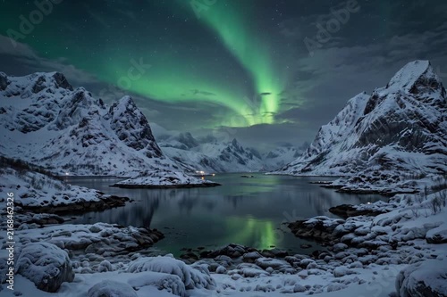 Stunning view of the Northern Lights aurora borealis shining in the night sky over snow-covered mountains and reflecting in the calm waters of a frozen fjord in winter