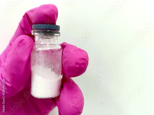 A gloved hand holds a small, clear glass medical vial containing freeze-dried white powder.