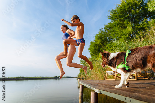 Mother and son jumping together into lake water while dog watches from wooden dock.