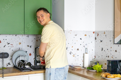Bild auf Leinwand Man washes dishes and turns around smiling while doing household chores in modern kitchen