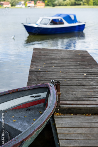 Weathered rowboat moored beside a wooden dock, with a blue motorboat softly blurred on the calm lake in the background.