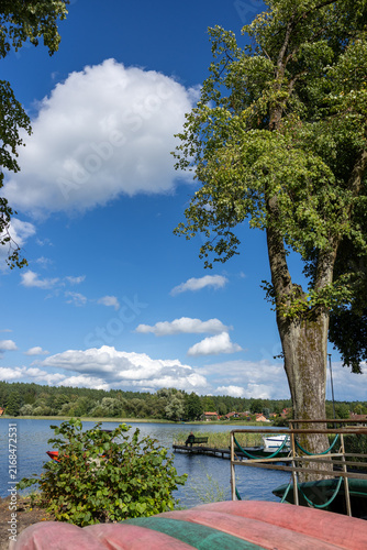 Solitary person sitting on a bench at the end of a narrow wooden pier, looking across a peaceful lake toward a rural village under white summer clouds.