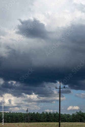 Massive dark storm cloud hanging above power poles, open fields and a distant pine forest, creating an ominous rural weather scene.