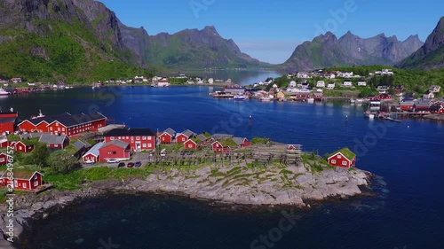 Aerial view gliding over traditional red rorbu cabins, drying racks, and rocky coastline surrounded by fjords and mountains in Reine, Lofoten Islands, Norway.