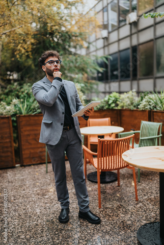 Obraz na plátně A young businessman stands outside holding a tablet and looking thoughtful