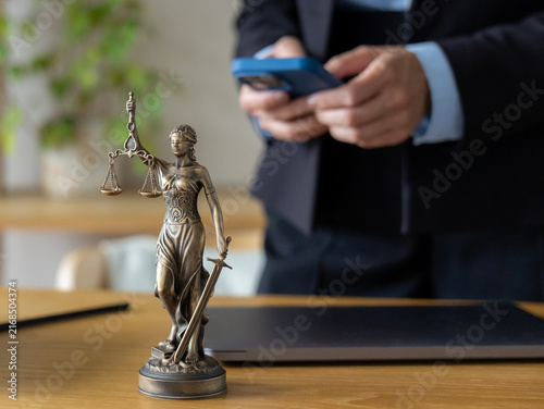 Bronze Lady Justice statue with scales sword. Blurred lawyer in suit uses blue smartphone behind. Wooden desk, tablet nearby. Modern online legal consultation and internet attorney services concept.