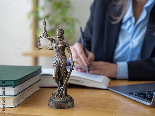 Professional internet lawyer providing online legal consultation service. Lawyer writing notes in open notebook with pen. Bronze Lady Justice statue stands on wooden desk near green books. 