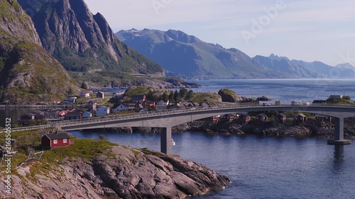 Aerial view of a coastal road bridge connecting rocky islands and traditional rorbu cabins in Reine, Lofoten, Norway on a clear day.