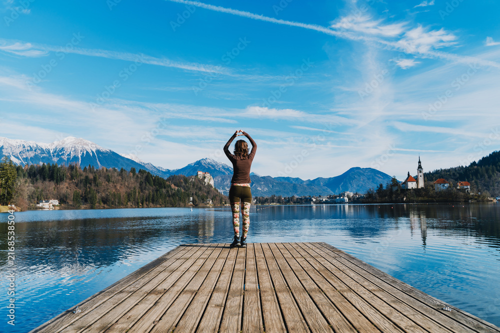 Fototapeta premium A young woman makes a heart gesture with her hands on a wooden pier at Lake Bled, Slovenia. Joyful travel lifestyle concept captures love, positivity, and freedom in scenic nature.