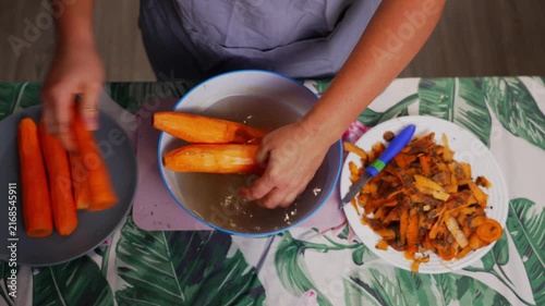 A person peeling carrots peeling carrots with a knife, in a kitchen countertop with a green leafy patterned, with carrots, knife, plate of food, with wooden countertop, ceramic plate texture.