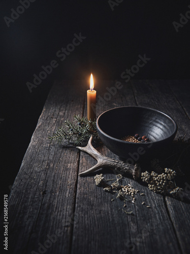Lit candle beside dark ceramic bowl with seeds on rustic wooden table, mystical altar still life