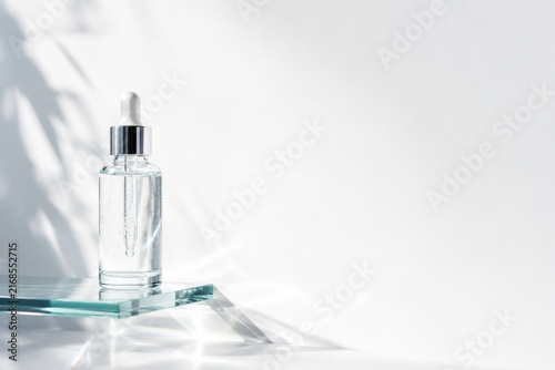A clear glass dropper bottle with liquid inside is placed on a transparent surface, casting delicate shadows on a bright white background.
