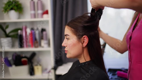 A hair stylist is professionally styling a client's hair in a salon setting, captured from a side view. The scene highlights hair products and shelves with bottles, set against a pink shirt.
