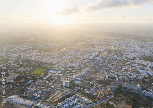 Aerial view of a residential town with scattered buildings, green spaces, and roads under a warm sunset light in Portugal.