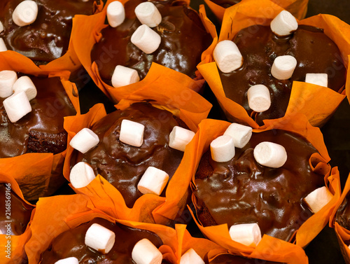 Cupcakes covered with dark chocolate and marshmallow pieces in orange paper wrappers displayed in a bakery, showcasing fresh sweet pastries for customers