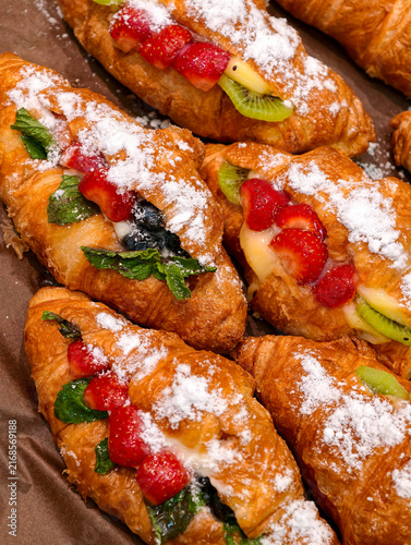 Fresh fruit filled croissants with strawberry kiwi and blueberries displayed in a bakery, highlighting sweet baked pastries finished with powdered sugar for customers