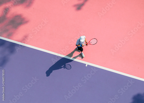 aerial view of a young tennis player in action on a vibrant pink and purple hard court. Dappled tree shadows and strong sunlight create a dynamic and aesthetic summer sports composition.