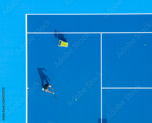 aerial view of a tennis player preparing to serve on a bright blue hard court. Long dramatic shadows, crisp white lines, and a basket of yellow balls create a striking sports composition.