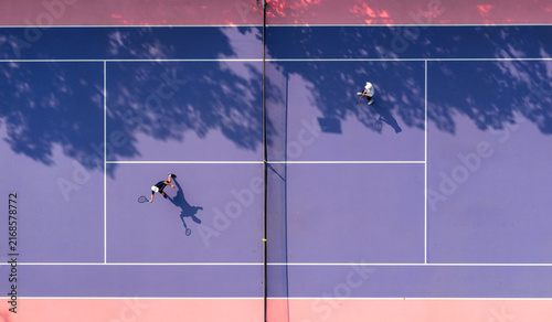 aerial view of young tennis players in action on a vibrant pink and purple hard court. Dappled tree shadows and strong sunlight create a dynamic and aesthetic summer sports composition.