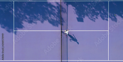 aerial view of a young tennis player in action on a vibrant pink and purple hard court. Dappled tree shadows and strong sunlight create a dynamic and aesthetic summer sports composition.